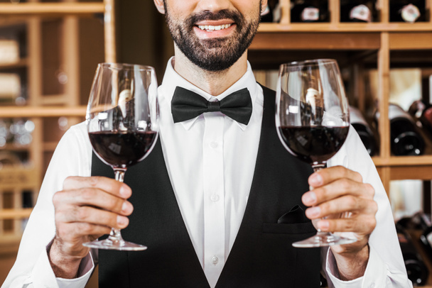 cropped shot of smiling young sommelier holding two glasses of red wine at wine store - Fotó, kép
