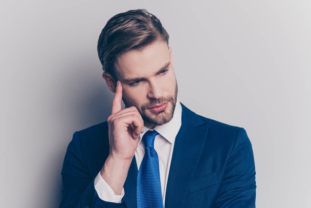 Portrait of thoughtful, smart, attractive, stunning man with stubble, modern hairstyle in blue suit touching temple with forefinger looking to the side isolated on grey background - Photo, Image