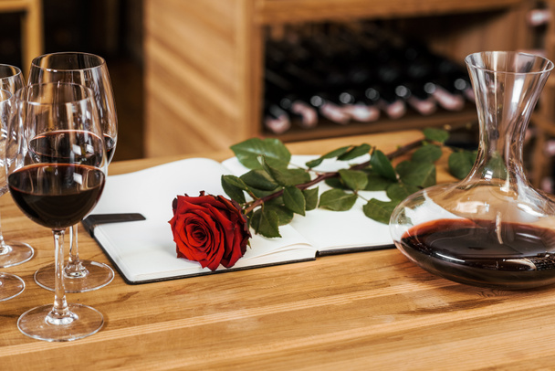 Detailní záběr karafa červeného vína s Poznámkový blok a rudá růže na dřevěný stůl na skladování vína - Fotografie, Obrázek