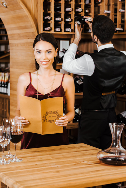 uśmiechnięta kobieta, posiadających karty menu przechowywania wina podczas steward biorąc butelkę z półki na tle - Zdjęcie, obraz