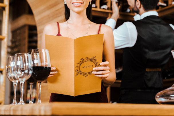 recortado disparo de la mujer sonriente celebración de la tarjeta de menú en la tienda de vinos mientras mayordomo tomar botella de estante en el fondo
 - Foto, Imagen