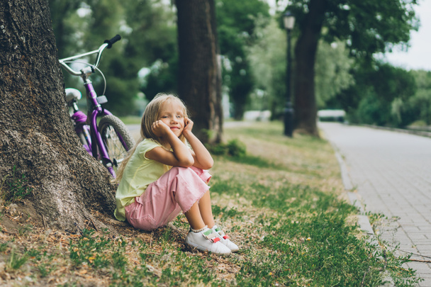 vista lateral da criança pensativa que descansa perto da bicicleta sob a árvore no parque
 - Foto, Imagem