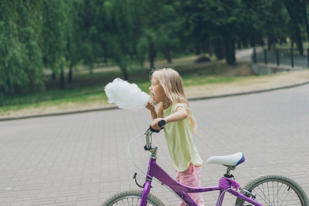 side view of adorable kid with bicycle eating cotton candy in park - Fotografie, Obrázek