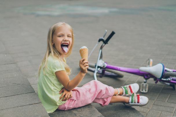 cute child with ice cream sticking tongue out while sitting on city steps alone - Fotografie, Obrázek