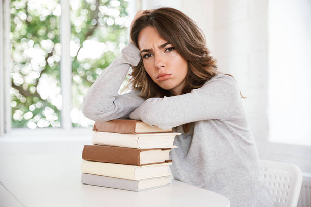 Image of sad displeased tired young lady sitting in classroom holding hands on a lot of books. - 写真・画像