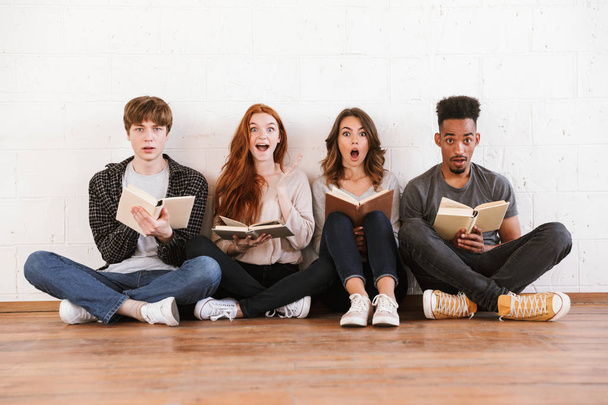 Image of shocked emotional young friends students sitting over white wall backgound indoors looking camera reading books. - Photo, image