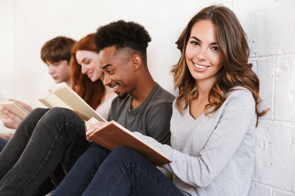 Imagem de jovens amigos estudantes sentados sobre backgound parede branca dentro de casa lendo livros. Concentre-se na mulher alegre
. - Foto, Imagem