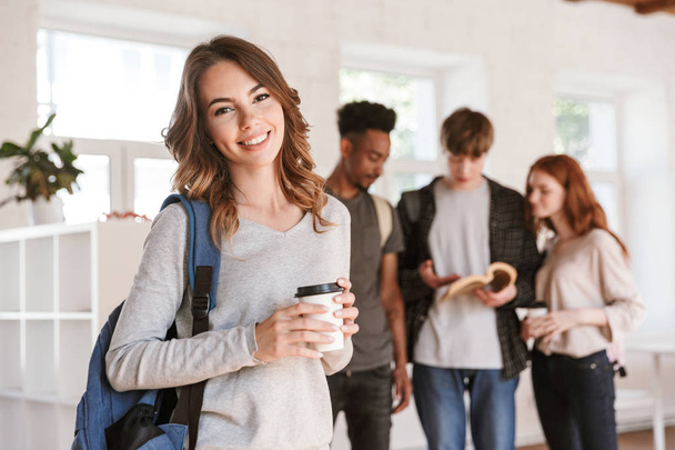 Image of young cheerful friends students standing in classroom indoors talking with each other. Focus on happy woman drinking coffee. - Foto, Imagem