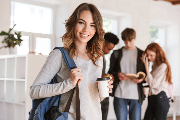 Image of young cheerful friends students standing in classroom indoors talking with each other. Focus on happy woman drinking coffee. - Foto, immagini