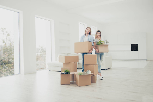 Dream of all our life! Buyer mover owner belongings concept. Two charming cheerful with toothy smile people holding carrying packages with thinks in hands standing in spacious light white living room - Fotó, kép