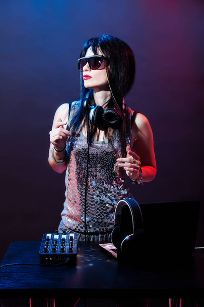 Retrato de DJ mujer joven sobre fondo rojo y azul. DJ mujer con headphne y gafas negras juega música disko. Fiesta de glamour en el equipo en el club de verano
. - Foto, Imagen