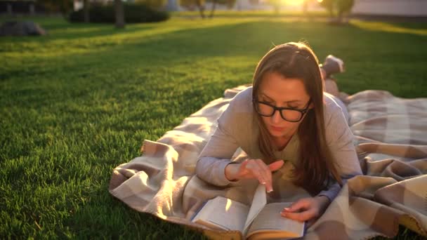 Girl in glasses reading book lying down on a blanket in the park at sunset - Filmagem, Vídeo