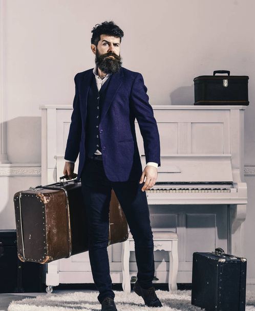 Macho stylish on strict face stands and carries big vintage suitcase. Baggage and travelling concept. Man, traveller with beard and mustache with baggage, luxury white interior background - Photo, Image