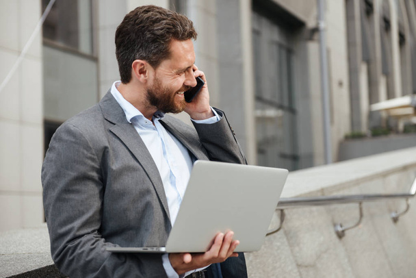 Photo of happy businesslike man 40s in gray suit working on silver laptop and talking on mobile phone while standing near business center - Photo, Image