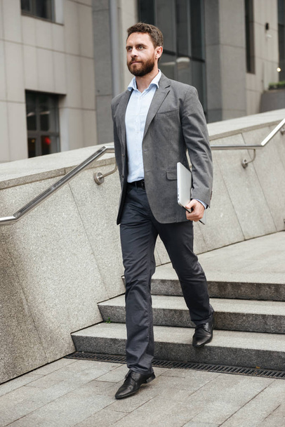 Full length photo of mature business man in formal wear holding silver laptop and walking in front of office building in downtown - 写真・画像