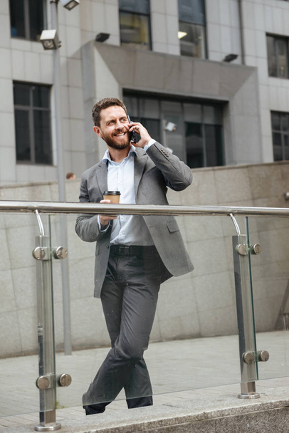 Full length photo of mature man in gray suit looking aside with smile while standing and drinking takeaway coffee in front of modern business center during business call - Foto, afbeelding
