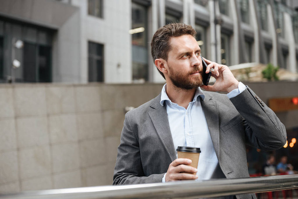 Photo of mature man in gray suit looking aside while standing and holding takeaway coffee in front of modern business center during business call - Foto, afbeelding
