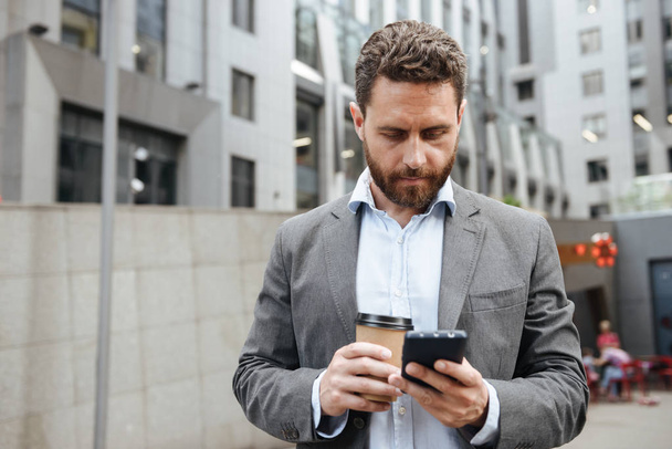Photo of adult man in gray suit looking at mobile phone in hand while standing with takeaway coffee in front of modern business center - Foto, afbeelding