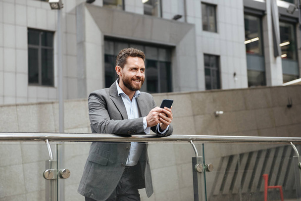 Photo of adult man in gray suit typing or reading text message in mobile phone while standing in front of modern office building - Zdjęcie, obraz