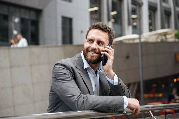 Portrait closeup of smiling joyful business man 40s in gray suit speaking on mobile phone while standing near office building on background - Photo, image