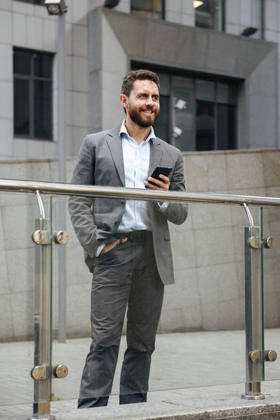 Full length photo of adult man in gray suit holding smartphone and looking ahead while standing in front of modern office building - Fotografie, Obrázek