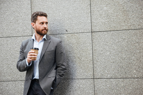 Photo closeup of adult concentrated man in gray suit and white shirt standing against granite wall and looking aside at copyspace with takeaway coffee in hand - Fotografie, Obrázek