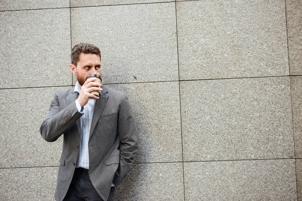 Photo closeup of adult handsome man in gray suit and white shirt standing against granite wall and looking aside at copyspace while drinking takeaway coffee - Foto, imagen