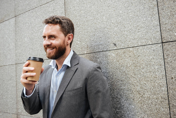 Portrait closeup of adult handsome man in gray suit and white shirt standing against granite wall and smiling while drinking takeaway coffee - Photo, image