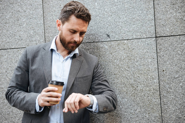 Photo closeup of adult businessman in gray suit and white shirt standing against granite wall and looking at wrist watch while drinking takeaway coffee - Fotografie, Obrázek