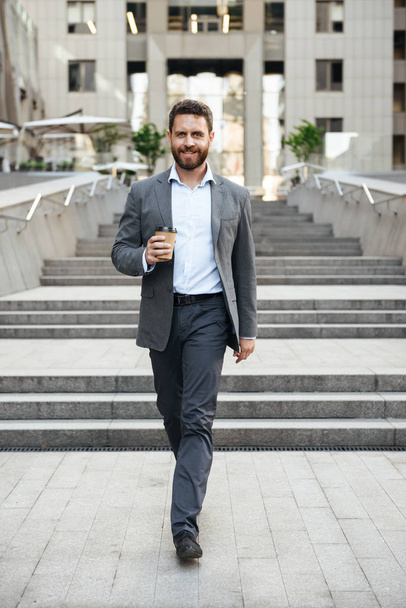 Photo of pleased entrepreneur man 40s in gray suit walking down stairs of modern business center with takeaway coffee in hand - Фото, зображення