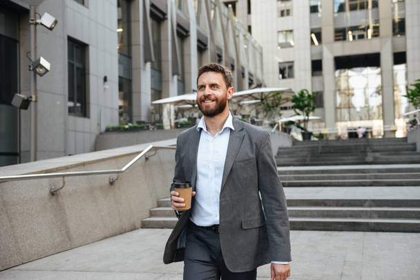 Photo closeup of successful happy man 40s in gray suit holding takeaway coffee in hand and walking down stairs of modern business center in downtown - Foto, Imagen