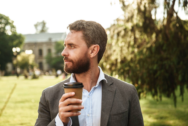 Photo closeup of adult handsome man in gray suit and white shirt looking aside while drinking takeaway coffee during walk in green park on sunny day - Фото, зображення