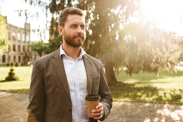 Magabiztos férfi 40-es szürke klasszikus ruha, séta gyönyörű park elvihető kávéval a kezében, a napos-fotó - Fotó, kép
