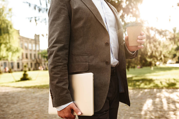 Cropped photo of adult man in gray classical suit walking through park carrying takeaway coffee and silver laptop - Foto, Bild