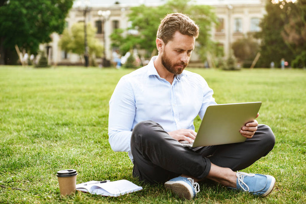 Photo of concentrated caucasian man in business clothing sitting on grass in park with legs crossed while working on silver laptop - Fotoğraf, Görsel