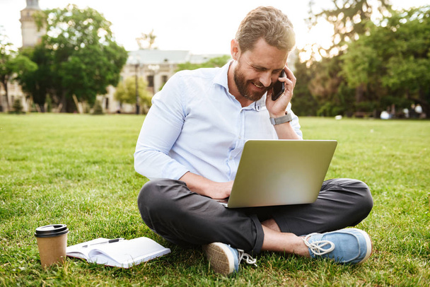 Photo of adult european man in business clothing sitting on grass in park with legs crossed and having business call while working on silver laptop - Photo, image