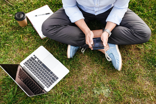 Cropped photo from top of caucasian man in business clothing sitting on grass in park while using cell phone and silver laptop - Foto, Imagem