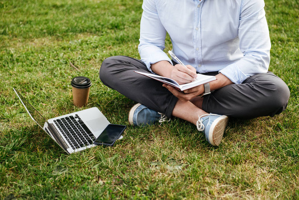 Cropped photo of caucasian man in business clothing sitting on grass in park with legs crossed and writing down notes in notebook while working on laptop - Fotó, kép