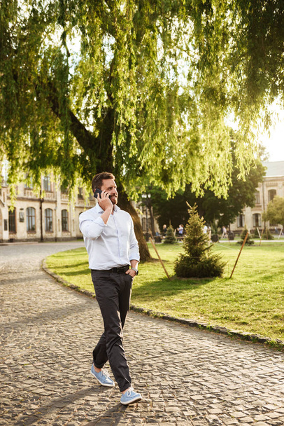 Full length photo of smiling businesslike man in formal wear walking through park on sunny day and talking on smartphone - Foto, Imagem