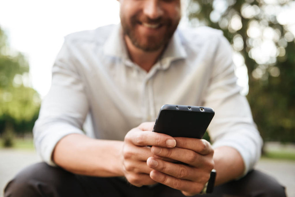 Rajattu kuva iloinen houkutteleva mies valkoinen paita istuu puistossa ja käyttää mustaa matkapuhelinta chat
 - Valokuva, kuva