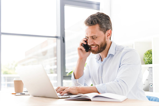 Image of happy handsome bearded man sitting in office working looking aside talking by mobile phone while using laptop computer. - Photo, Image