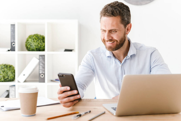 Image of happy handsome bearded man sitting in office working looking aside using mobile phone and laptop computer. - Photo, Image