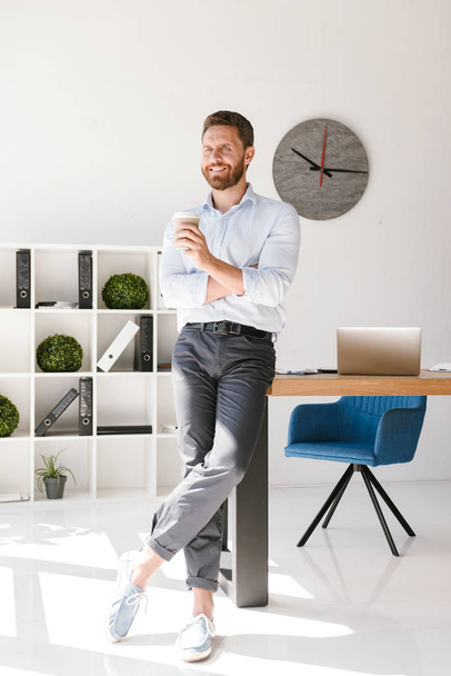 Attractive businessman drinking coffee while leaning on a desk in office - Valokuva, kuva
