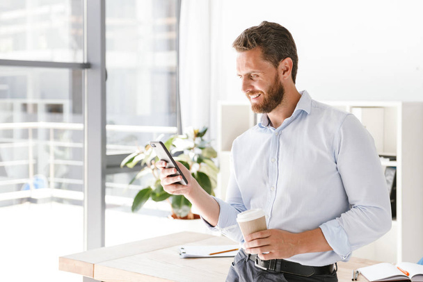 Happy businessman using mobile phone while drinking coffee in office - Foto, imagen