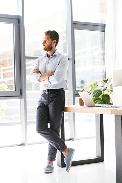 Image of handsome bearded man in office standing near table working looking aside. - Foto, Imagen