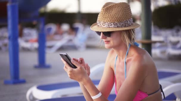 Woman in swimwear sitting by swimming pool surfing on smart phone - Filmati, video