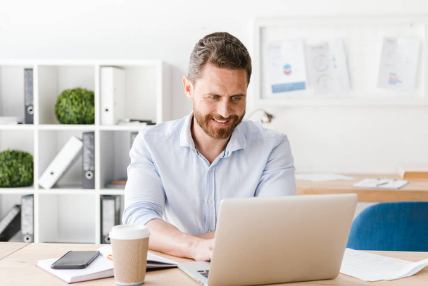 Image of happy bearded man sitting in office working on laptop computer. Looking aside. - Fotografie, Obrázek
