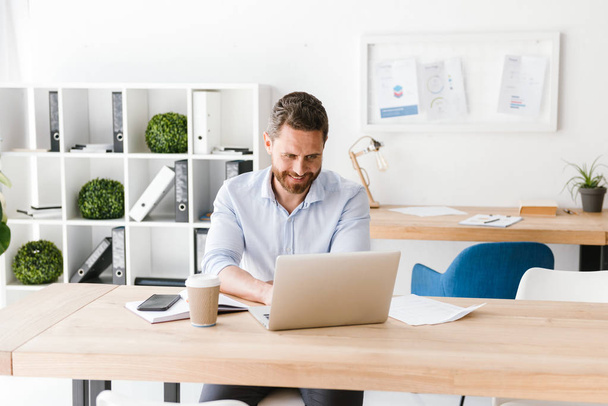 Image of happy bearded man sitting in office working on laptop computer. Looking aside. - Foto, Bild