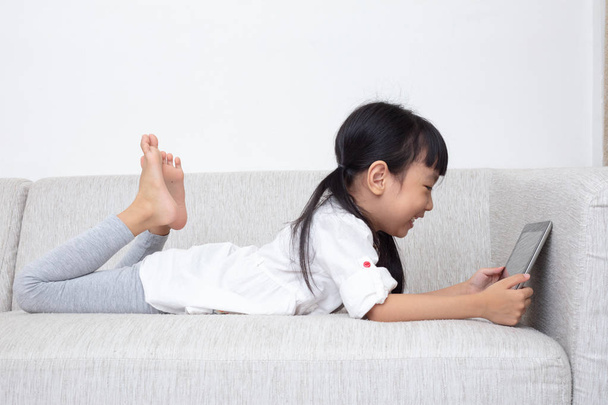 Asian Chinese little girl lying on the sofa and playing tablet in the living room - Foto, immagini