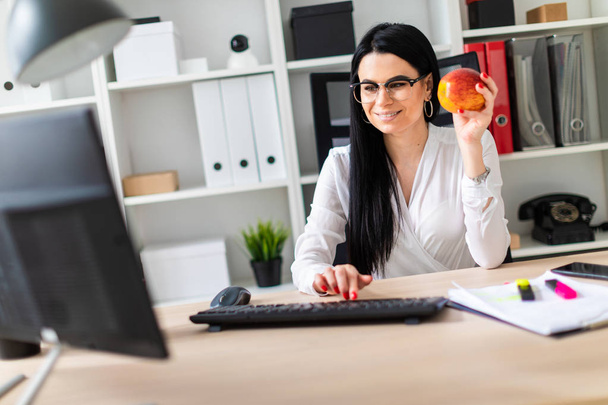 ein junges Mädchen sitzt an einem Computertisch, hält einen Apfel in der Hand und druckt auf der Tastatur. - Foto, Bild